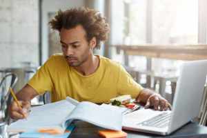 Inscrição para o FIES 2023: jovem negro sentado na mesa de frente para um notebook usa o computador para estudos.